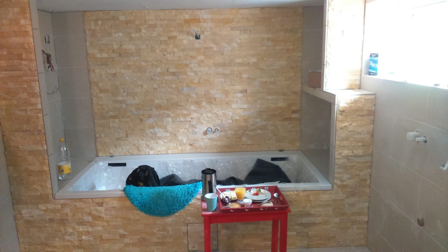 Łazienka w trakcie remontu: wanna zabezpieczona folią, ściany wyłożone dekoracyjną, beżową cegłą, na czerwonym stoliku śniadanie. Widoczne niedokończone prace instalacyjne.