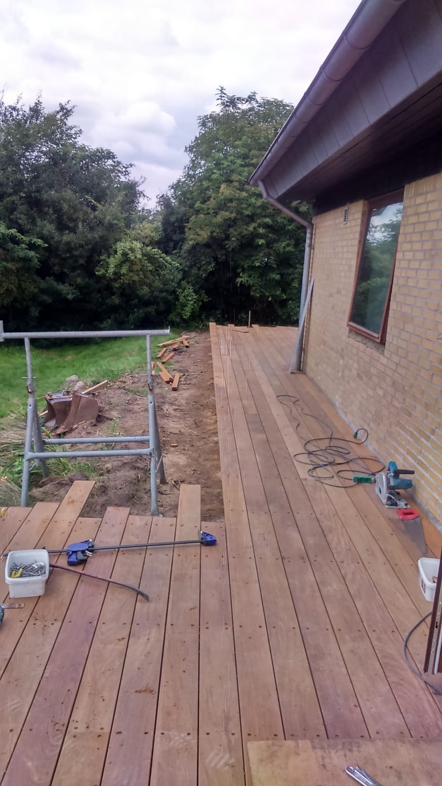 Budowa drewnianego tarasu przy domu z cegły, widoczny metalowy kozioł, narzędzia oraz deski tarasowe w trakcie montażu, zieleń drzew w tle.