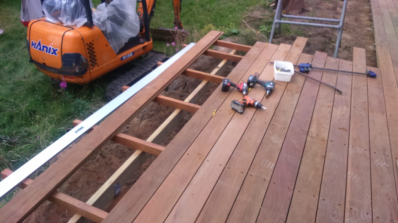 Budowa drewnianego tarasu: widoczny szkielet konstrukcji, częściowo ułożone deski, wkrętarki, skrzynka z wkrętami, mini koparka Hanix w tle.