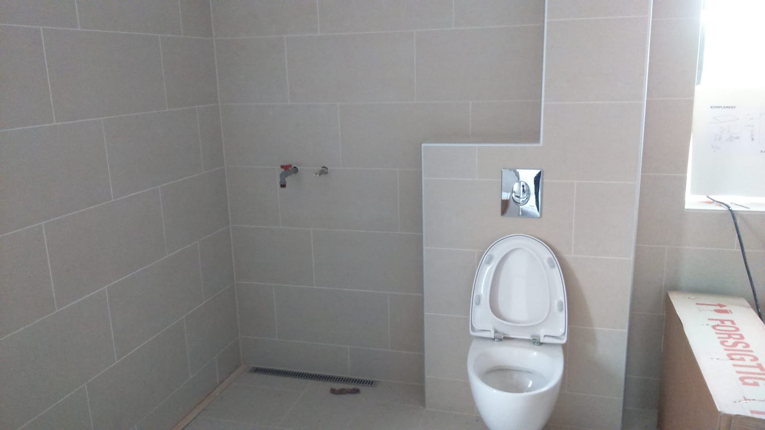 Wykończona łazienka z beżowymi płytkami, białą toaletą z otwartą klapą i chromowaną spłuczką, widoczny odpływ liniowy w podłodze i niezamontowana bateria na ścianie, obok okna karton z napisem.