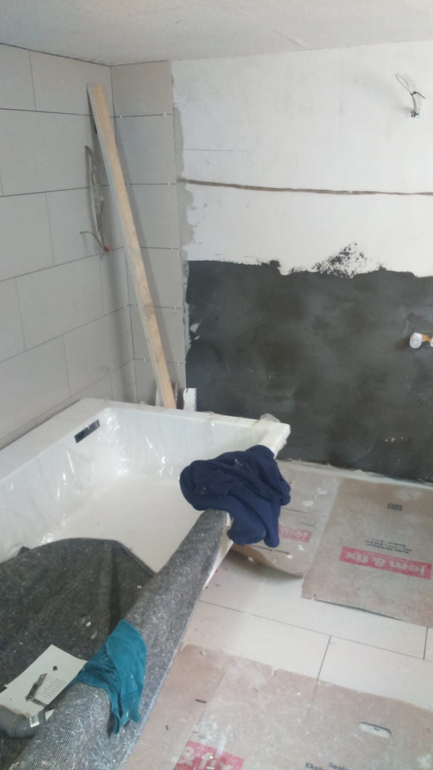 Remont łazienki w trakcie: częściowo wykafelkowana ściana, fragment ściany z nałożoną szarą masą, zabezpieczona folią wanna, podłoga wyłożona kartonem ochronnym.