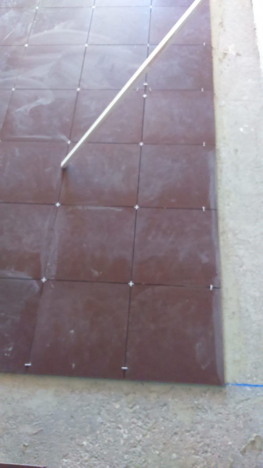 Układanie brązowych płytek podłogowych z krzyżykami dystansowymi, widoczna biała listwa poziomująca i surowy betonowy podkład.