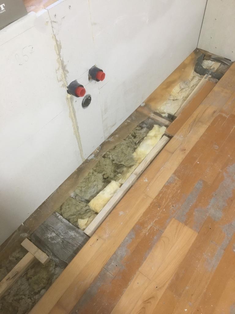 Fragment ściany z wystającymi rurami i niedokończoną podłogą z odsłoniętą izolacją, widoczne drewniane elementy konstrukcyjne i resztki materiałów budowlanych.