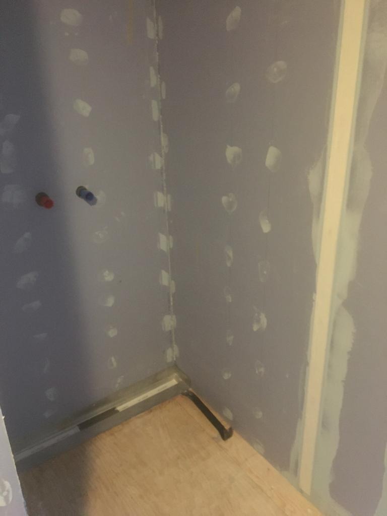 Ściana w trakcie szpachlowania z widocznymi punktowymi aplikacjami gipsu, przygotowana do dalszej obróbki, z widocznymi rurami instalacji wodnej i fragmentem podłogi.
