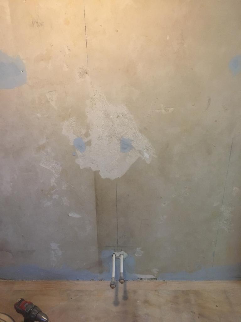Surowa ściana w trakcie remontu z widocznymi śladami po wcześniejszych pracach, częściowo pokryta szpachlą, z wystającymi rurami i wiertarką w rogu.