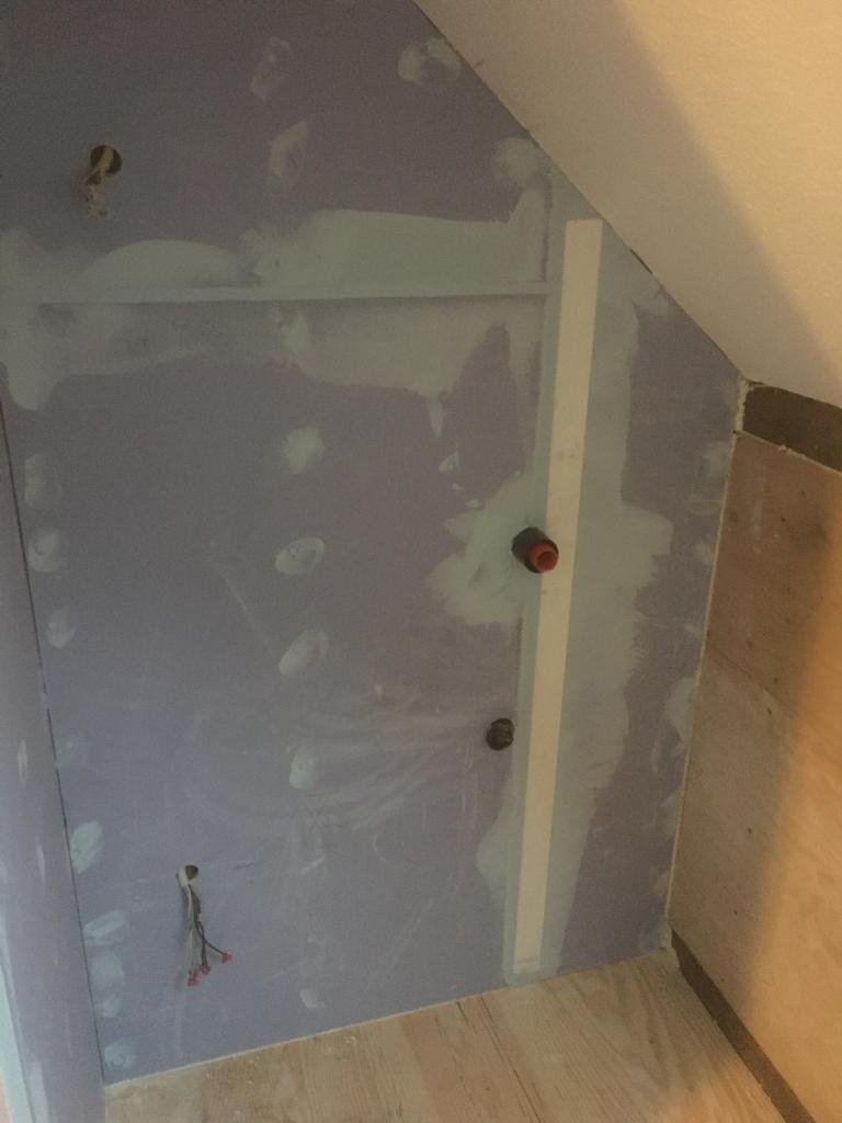 Ściana pod skosem w trakcie prac wykończeniowych, widoczne ślady szpachlowania i otwory na instalacje elektryczne, listwa przypodłogowa w trakcie montażu.