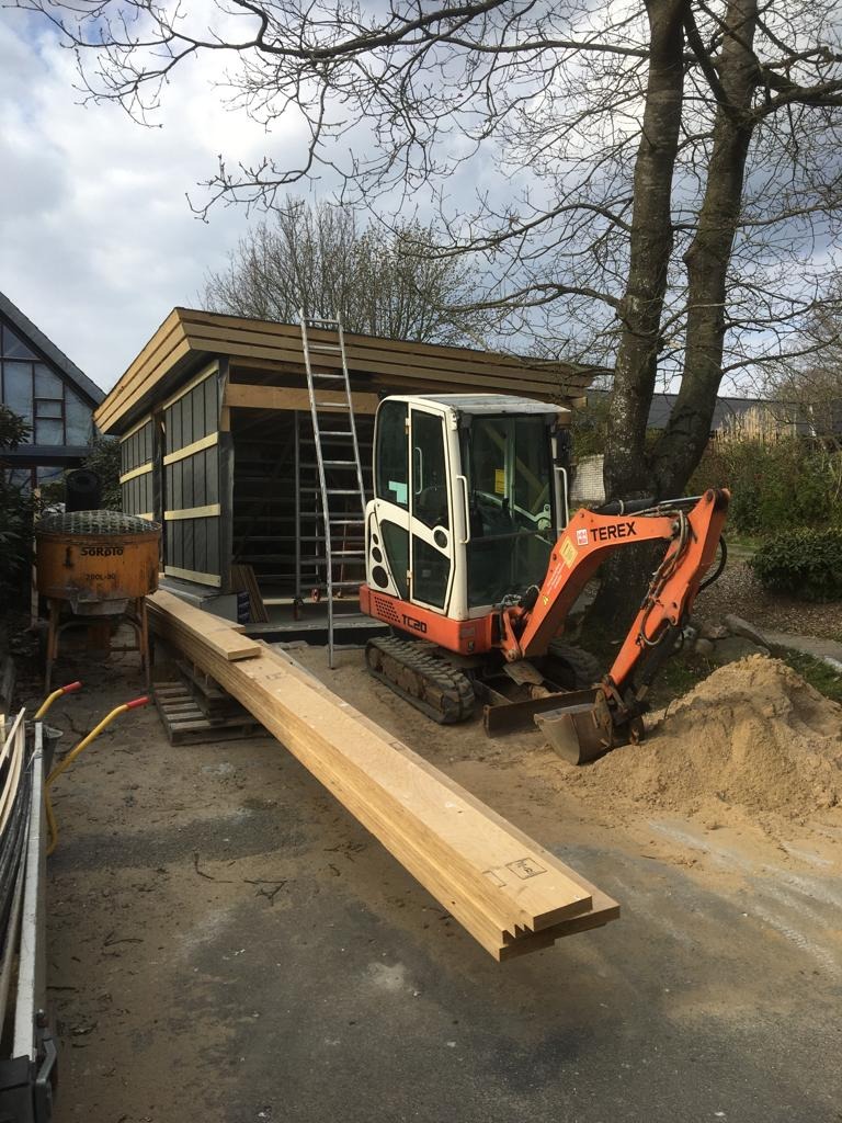 Wznoszona konstrukcja szkieletowa budynku gospodarczego z użyciem minikoparki Terex TC20, widoczne elementy drewniane, drabina oparta o konstrukcję oraz betoniarka w tle