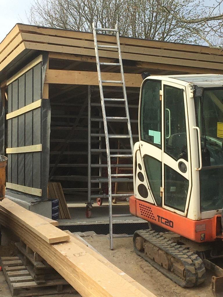 Budowa szopy lub małego budynku z widocznymi elementami konstrukcyjnymi, deskami, drabina aluminiowa i mała koparka TC20 obok.
