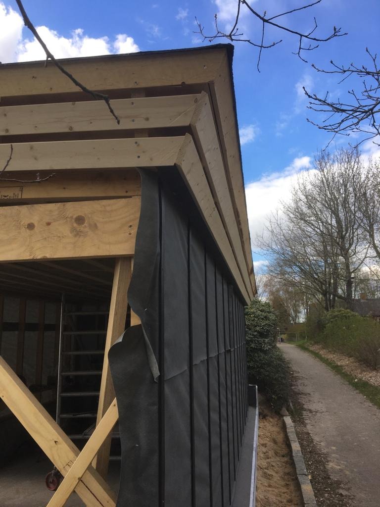 Konstrukcja drewniana z czarną membraną hydroizolacyjną na ścianie budynku w trakcie budowy, widoczny fragment dachu i niebo z chmurami.