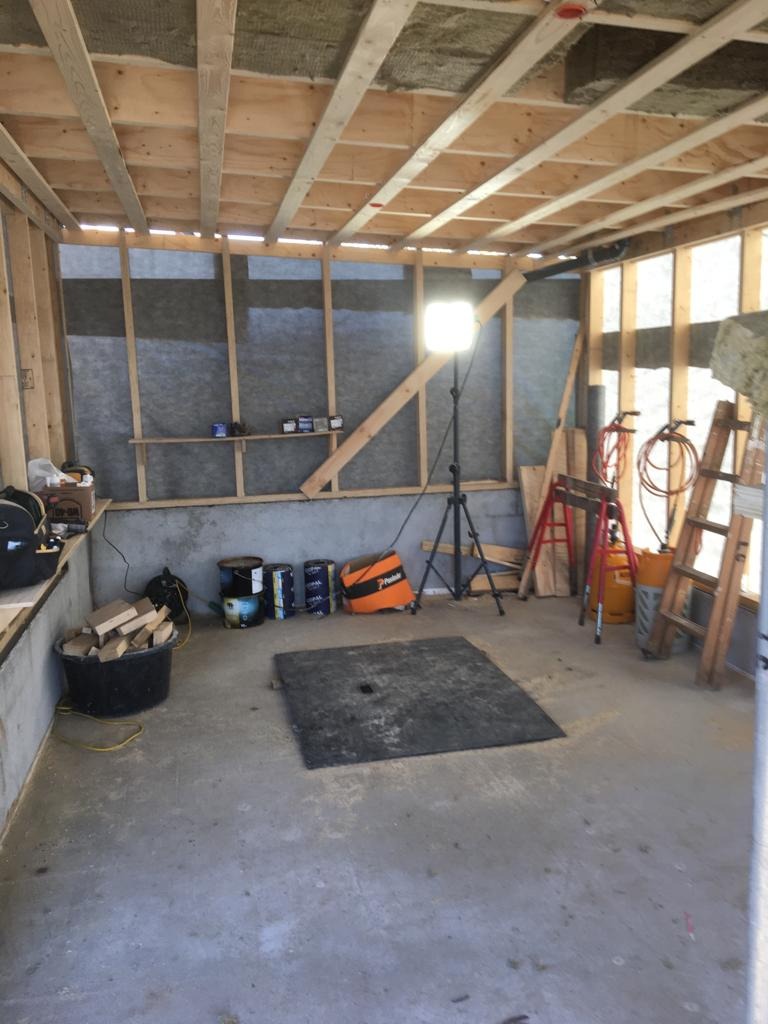 Wnętrze pomieszczenia w trakcie remontu, widoczne drewniane konstrukcje ścian i sufitu, ocieplenie ścian, puszki z farbą, wiadro z drewnem, lampa na statywie, drabiny i narzędzia.