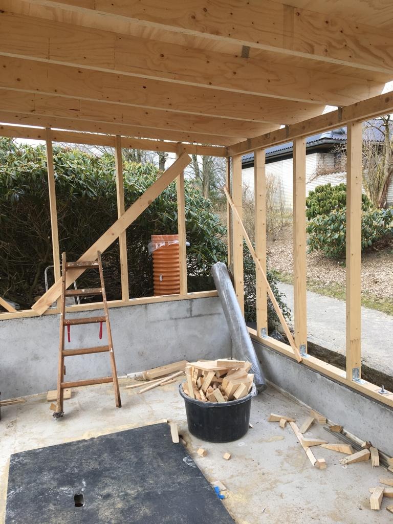 Surowa konstrukcja drewniana z widocznymi deskami, drabina oparta o ścianę, wiadro z kawałkami drewna i folia w tle, stan surowy budynku w trakcie budowy.