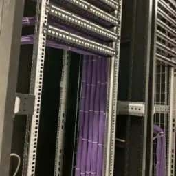 Widok z bliska na uporządkowane fioletowe kable sieciowe w szafie rack, prowadzone pionowo i zabezpieczone opaskami zaciskowymi, z widocznymi panelami krosowniczymi i metalową konstrukcją szafy.