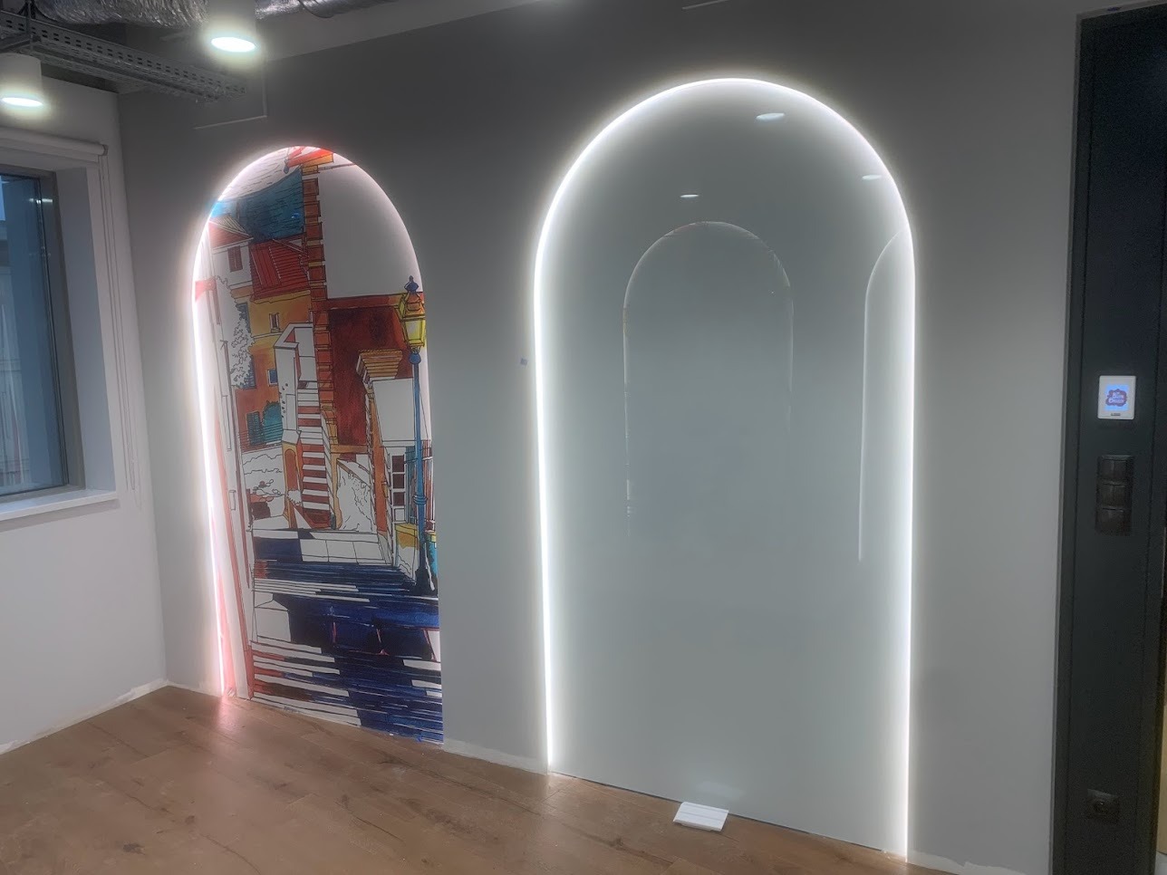 Nowoczesne wnętrze z dwoma podświetlanymi wnękami w kształcie łuku; jedna z grafiką przedstawiającą uliczkę miejską w stylu włoskim, druga pusta, z delikatnym odbiciem.