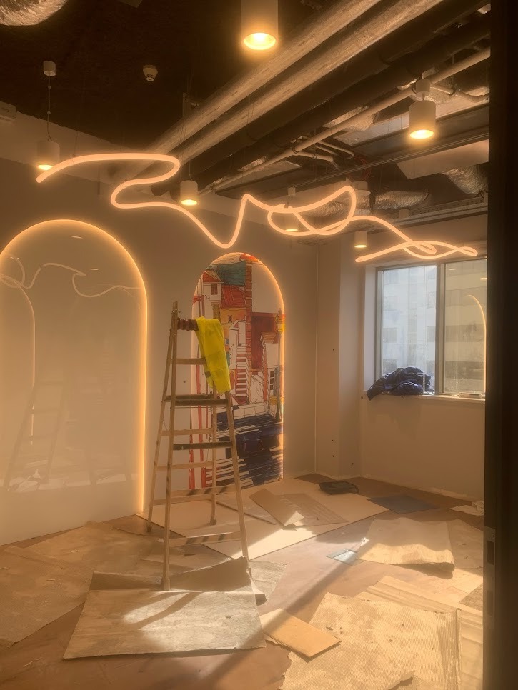 Pokój w trakcie prac wykończeniowych, widoczne resztki materiałów na podłodze, drabina oparta o ścianę z dekoracyjnym panelem, oraz neonowe oświetlenie sufitowe.