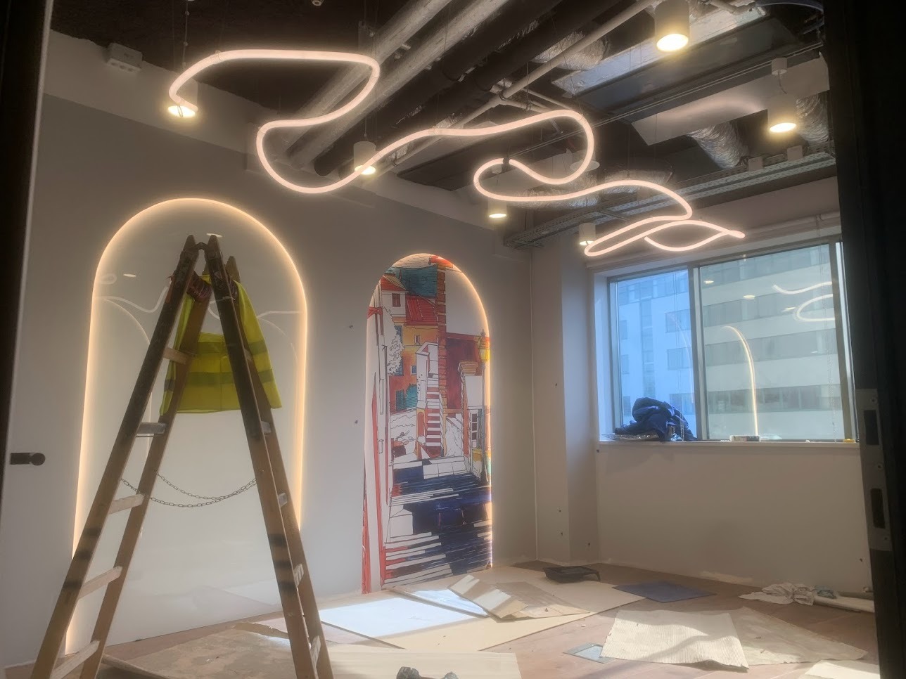 Wnętrze biurowe w trakcie prac wykończeniowych: drabina z żółtą kamizelką przy ścianie z podświetlanym łukiem, dekoracyjna lampa sufitowa w kształcie linii, okno z widokiem na budynek biurowy.