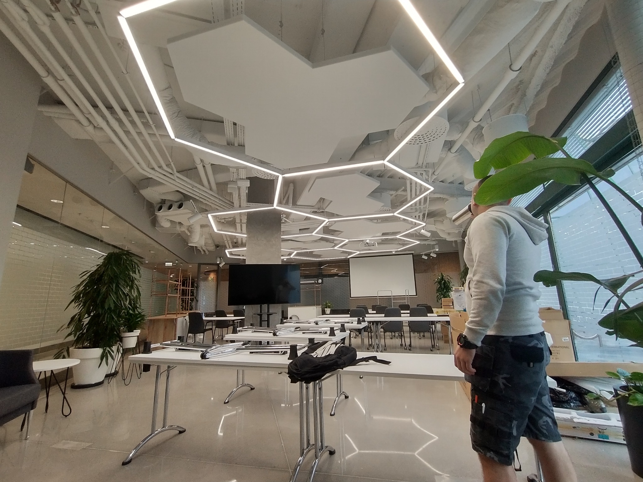 Nowoczesne biuro z geometrycznym oświetleniem LED na suficie, widok na instalacje pod sufitem, biurka i krzesła w tle, osoba w odzieży roboczej w prawym dolnym rogu.