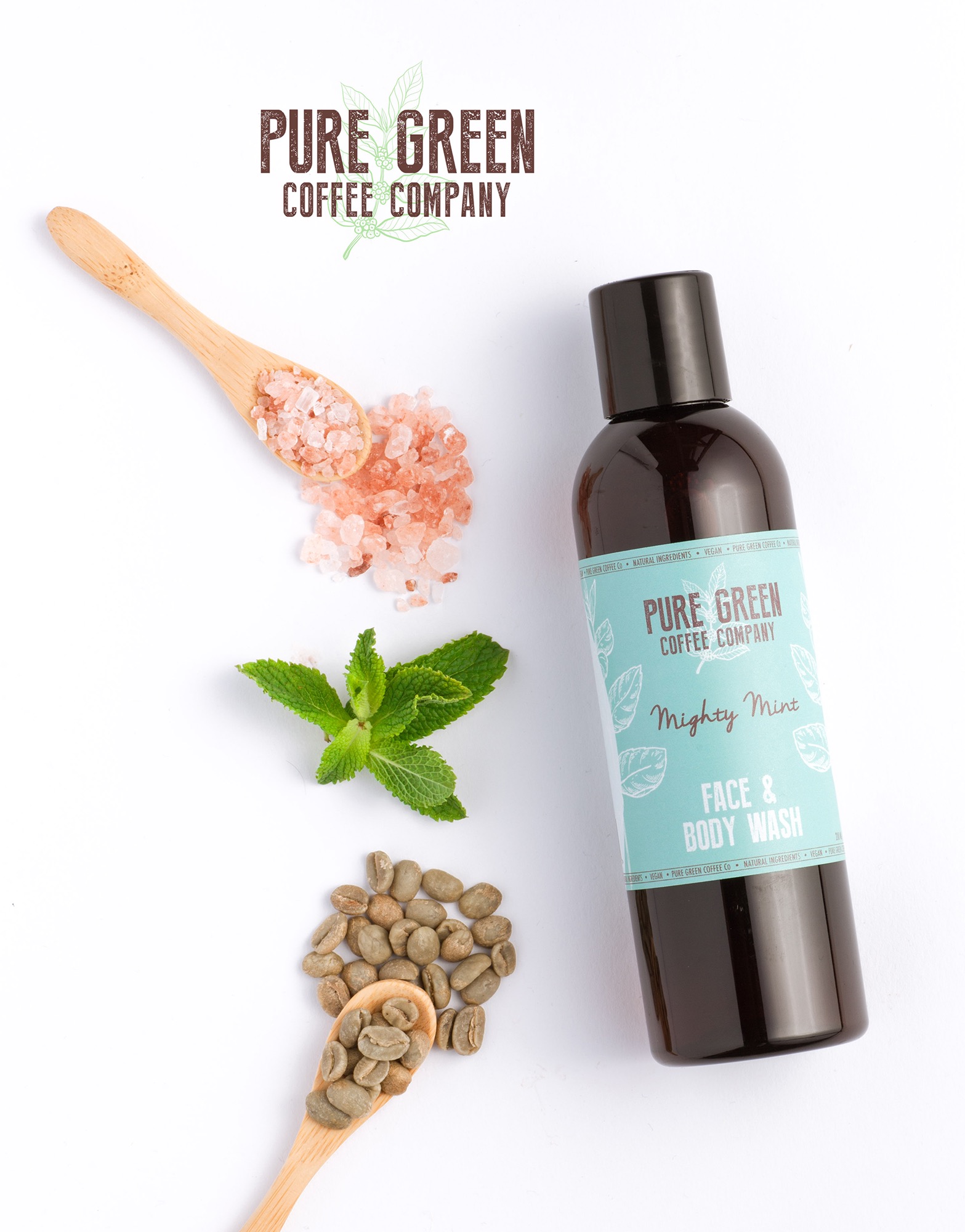 Butelka ciemnego płynu do mycia twarzy i ciała Mighty Mint marki Pure Green Coffee Company na białym tle z drewnianą łyżeczką z różową solą, gałązką mięty i drugą łyżeczką z zielonymi ziarnami kawy.