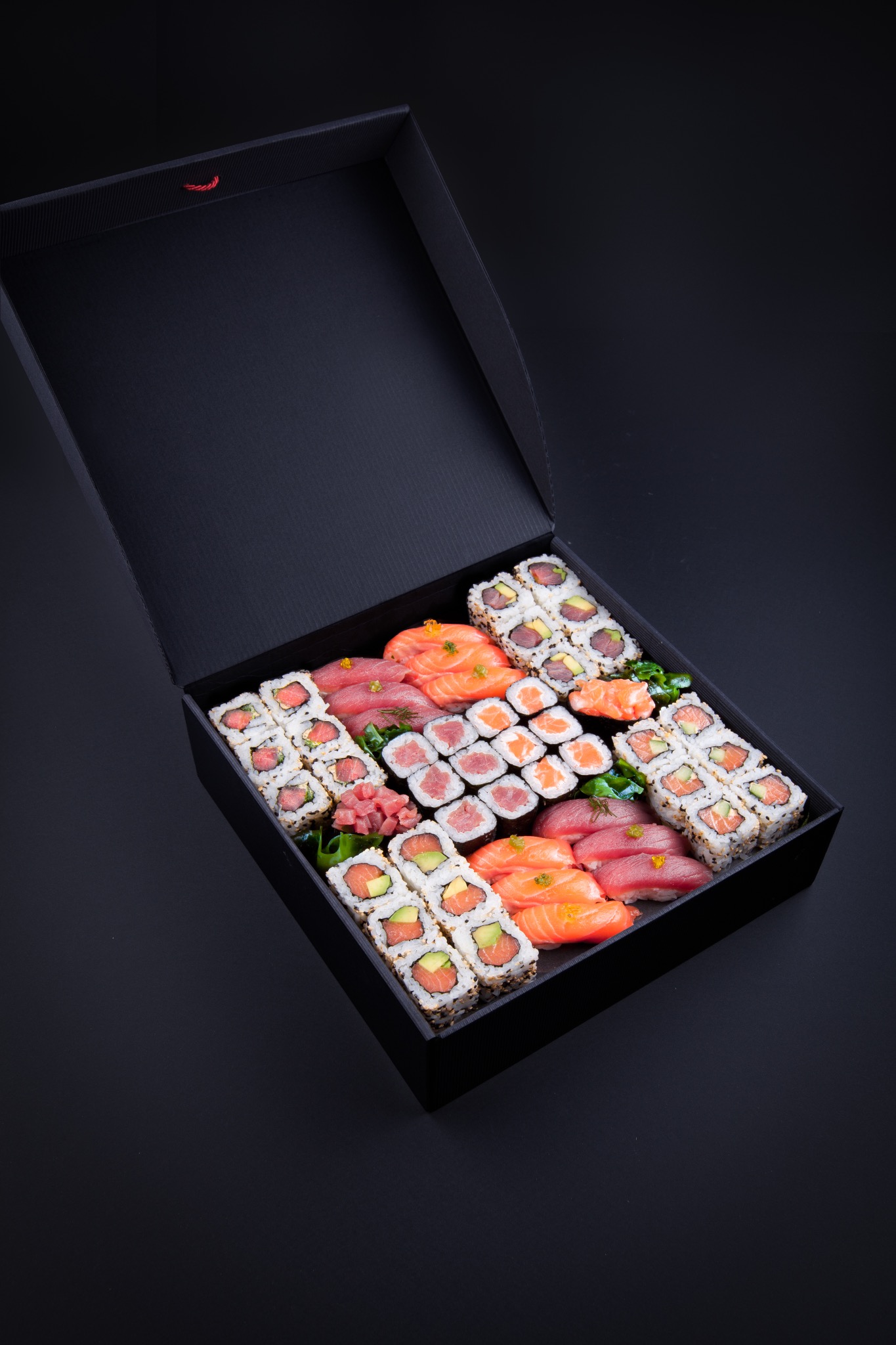 Zestaw sushi w eleganckim, otwartym czarnym pudełku z czerwonym sznurkiem, różnorodne rolki i nigiri ułożone na spodzie pudełka, na jednolitym czarnym tle.