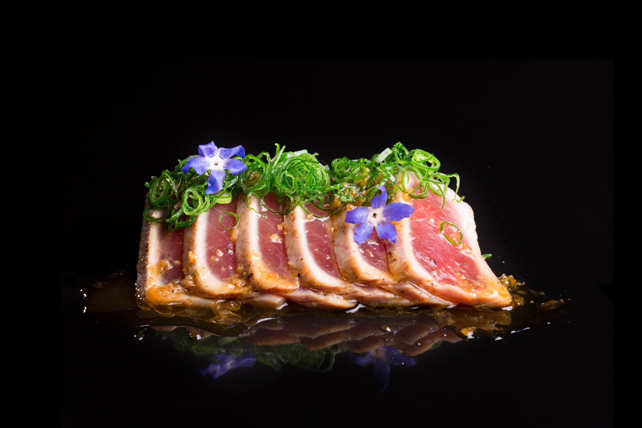 Plasterki grillowanego tuńczyka, udekorowane zielonym szczypiorkiem i fioletowymi kwiatami, ułożone na czarnej, błyszczącej powierzchni, tworzącej lustrzane odbicie.