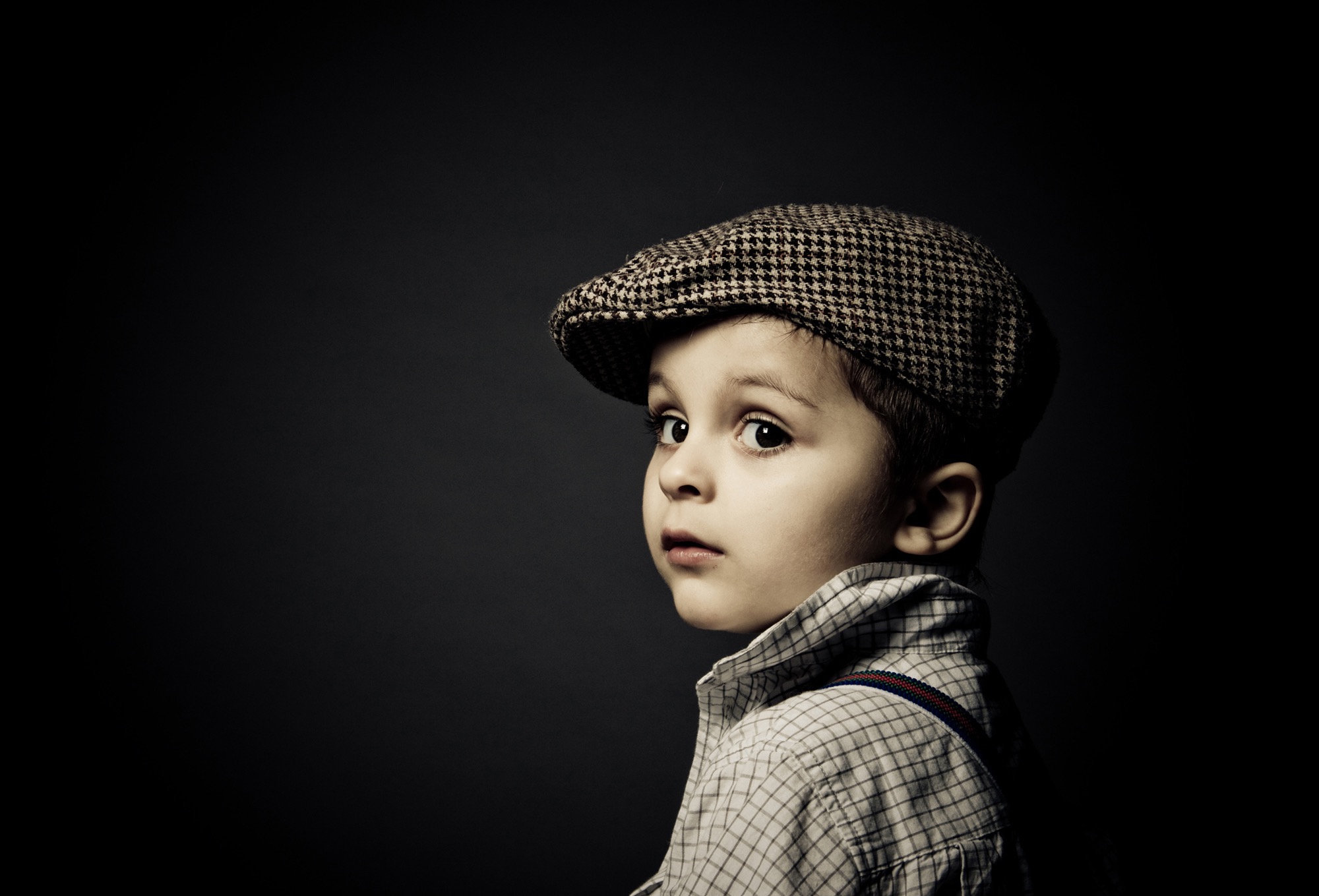Portret chłopca w kraciastej czapce i koszuli w kratę z szelkami, spoglądającego przez ramię na ciemnym tle.