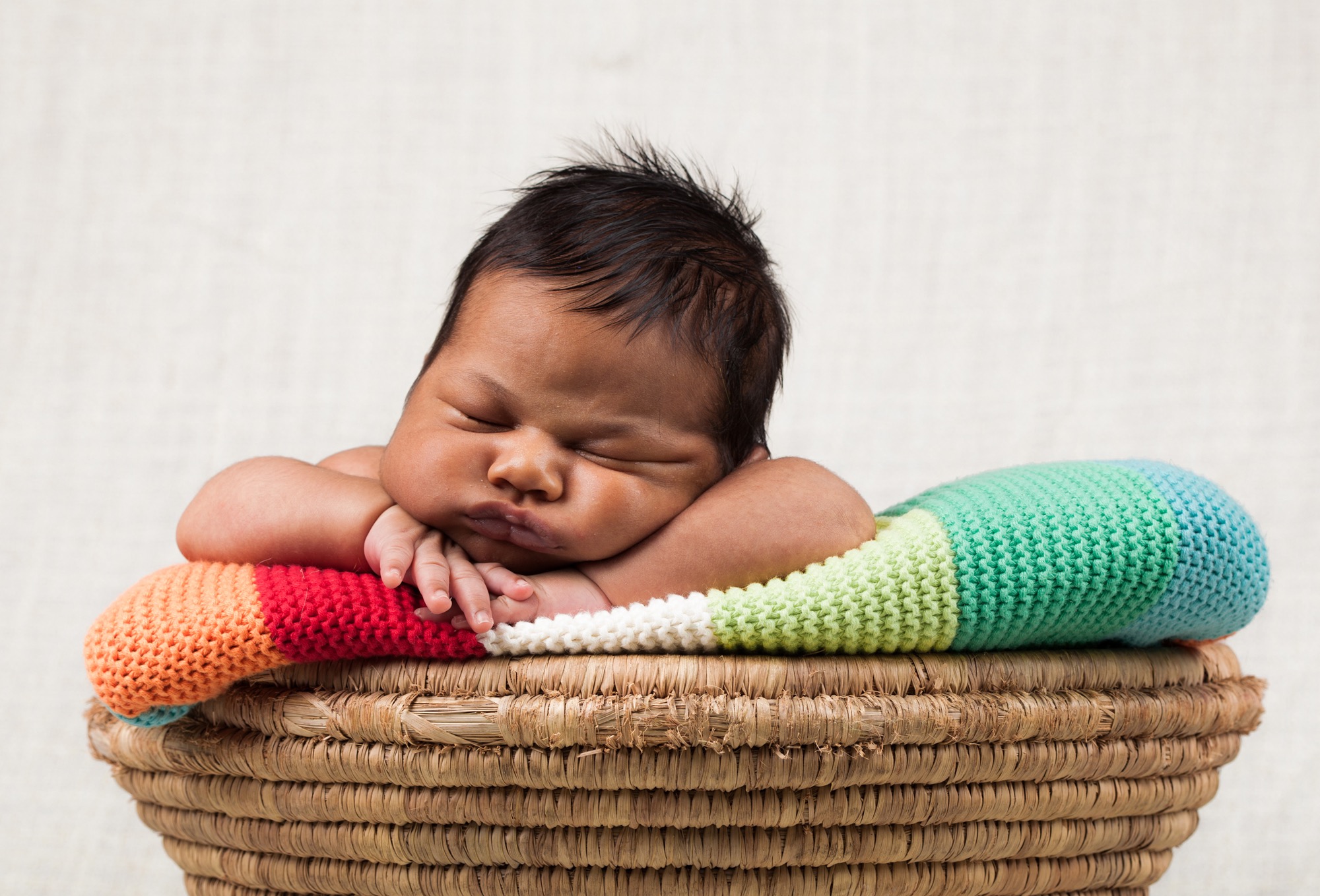 Śpiący noworodek z ciemną karnacją, ułożony w wiklinowym koszu wyściełanym kolorowym, dzierganym kocykiem w paski.