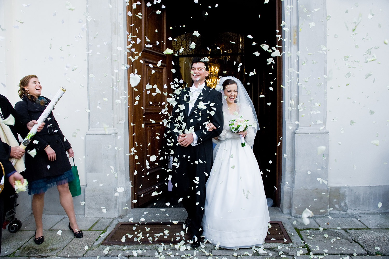 Uśmiechnięta para młoda wychodzi z kościoła obsypywana białymi płatkami kwiatów; w tle rozbawiona kobieta z tubą konfetti.