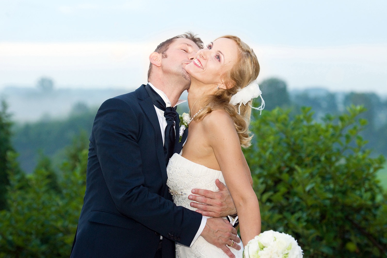 Panna młoda w sukni bez ramiączek, z bukietem białych kwiatów, obejmowana przez pana młodego w ciemnym garniturze, który całuje ją w policzek na tle zieleni i mglistego krajobrazu.
