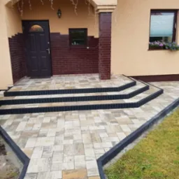 Nowo ułożona kostka brukowa przed domem z trzema stopniami i czarnym obrzeżem, widok na wejście i fragment ogrodu.