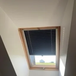 Ciemna przesłona okienna zamontowana w oknie dachowym z drewnianą ramą, widoczny fragment krajobrazu za oknem.