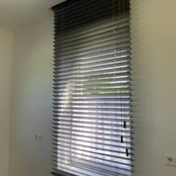 Zasłonięte, poziome, grafitowe żaluzje wewnętrzne z listwami o szerokości około 25mm, zamontowane w oknie z białymi ramami, widoczne gniazdka elektryczne na ścianie.