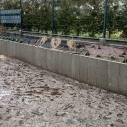 Betonowe ściany oporowe w ogrodzie w trakcie budowy, widoczne świeżo posadzone rośliny i mokry, błotnisty teren.