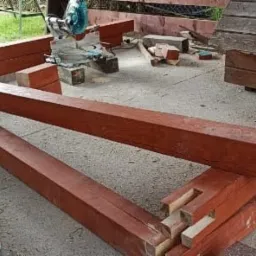 Konstrukcja z belek drewnianych z wycięciami, przygotowywana do montażu, z widoczną piłą, narzędziami i pojemnikami z klejem na szarym podłożu, w tle buda dla psa i trawnik.