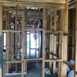 Wnętrze domu w trakcie budowy: drewniana konstrukcja szkieletowa ścian z widocznymi instalacjami, izolacją i betonową posadzką.