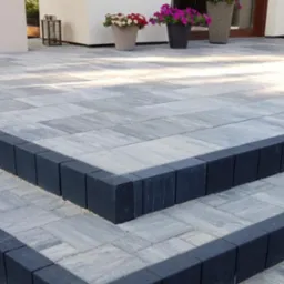 Schody wejściowe wyłożone szarą kostką brukową z ciemnymi obrzeżami, prowadzące do jasnego budynku z donicami z kwiatami.