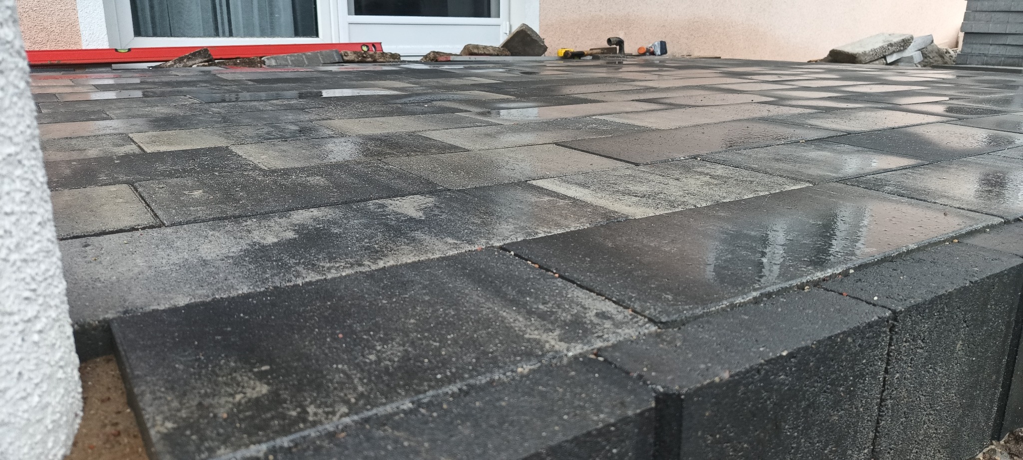 Świeżo ułożona, mokra, szara kostka brukowa na tarasie, widoczny poziomica, młotek i niedokończone krawędzie przy oknie.