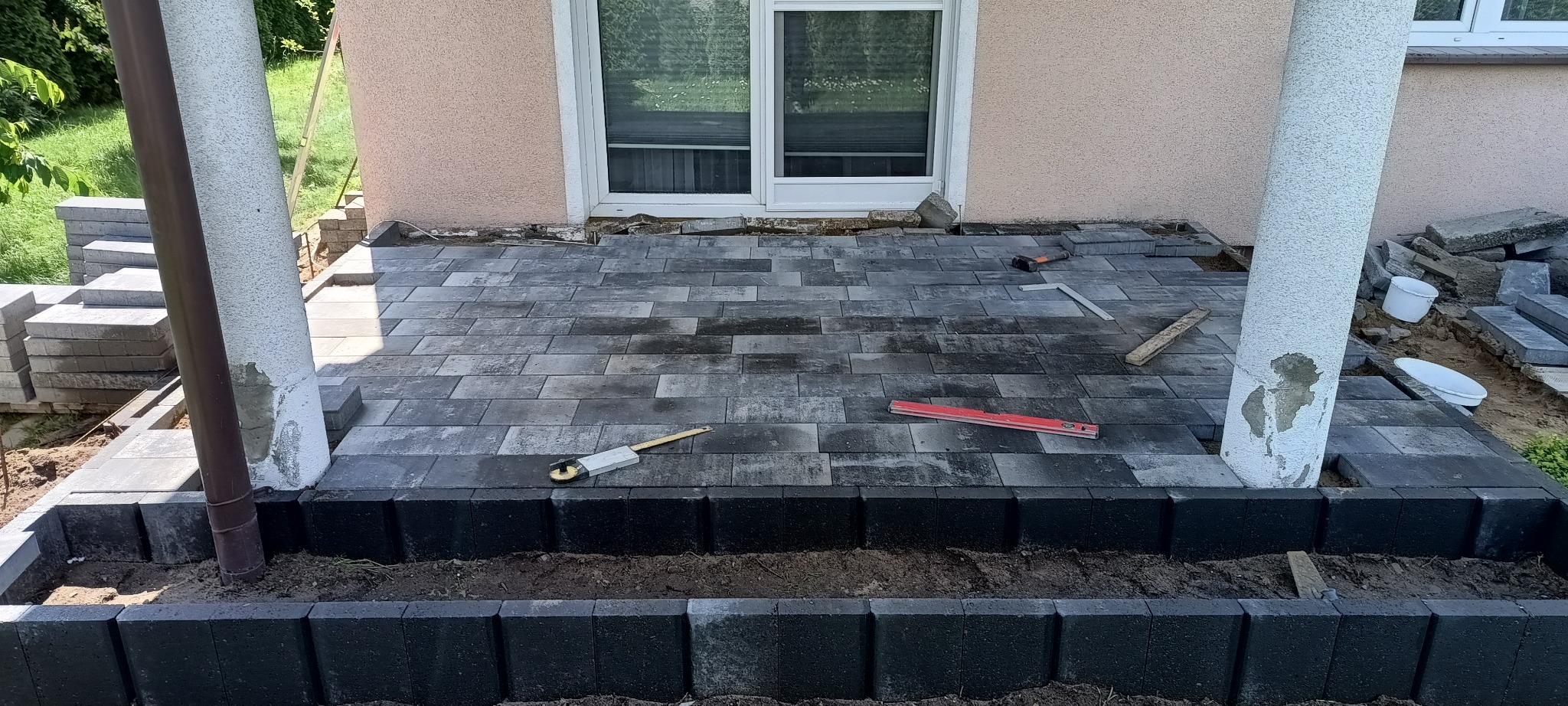 Fragment tarasu w trakcie budowy z szarej kostki brukowej, widoczne narzędzia pomiarowe, obrzeża z czarnej kostki, kolumny podtrzymujące zadaszenie, w tle okno budynku.