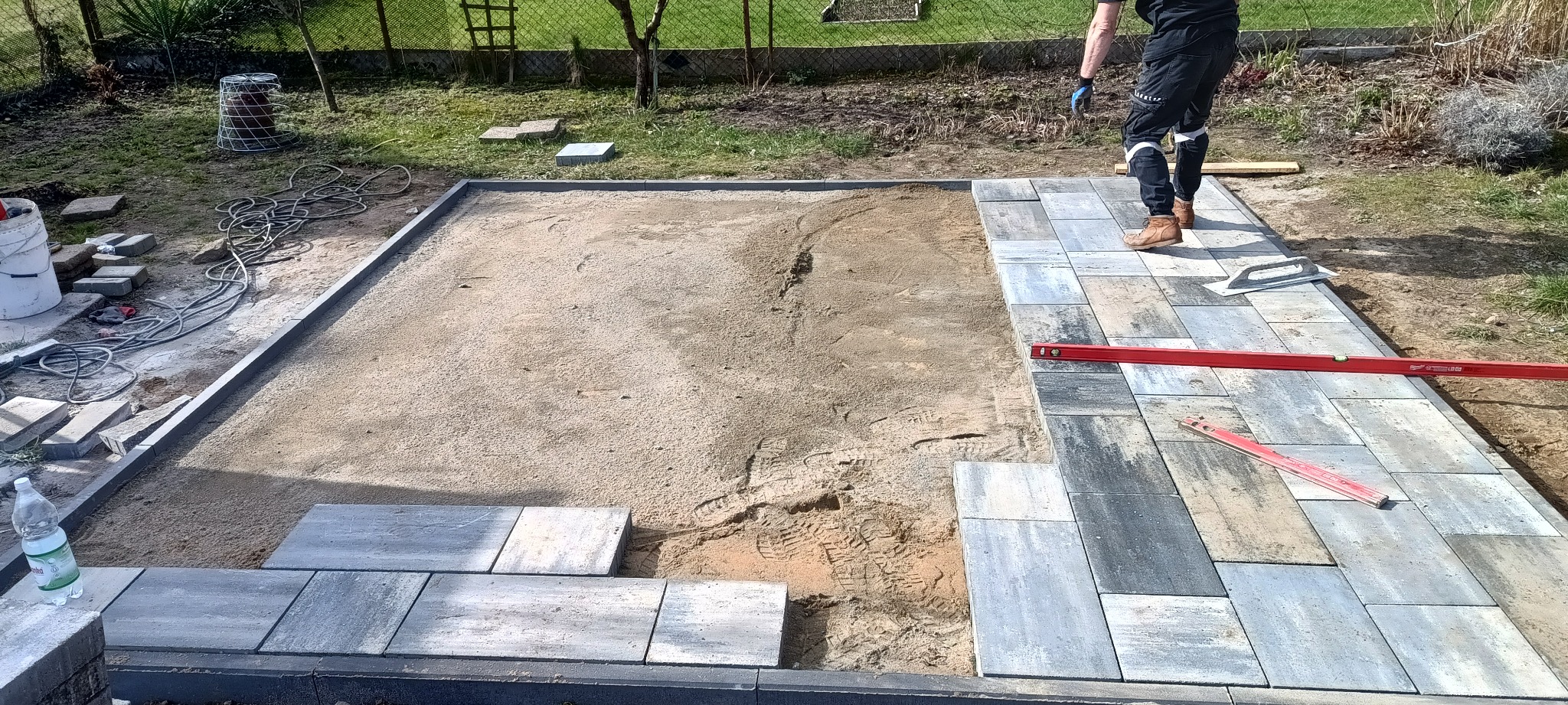 Prace brukarskie w toku: częściowo ułożona kostka brukowa w odcieniach szarości i błękitu, poziomica, piaskowe podłoże, widoczne ślady butów, pracownik w roboczych spodniach i brązowych butach...