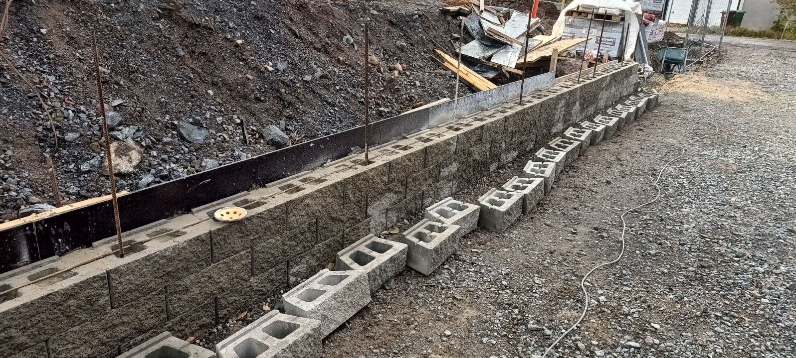 Wznoszenie muru oporowego z betonowych bloczków, z widocznym zbrojeniem pionowym i deskowaniem, na nierównym terenie.