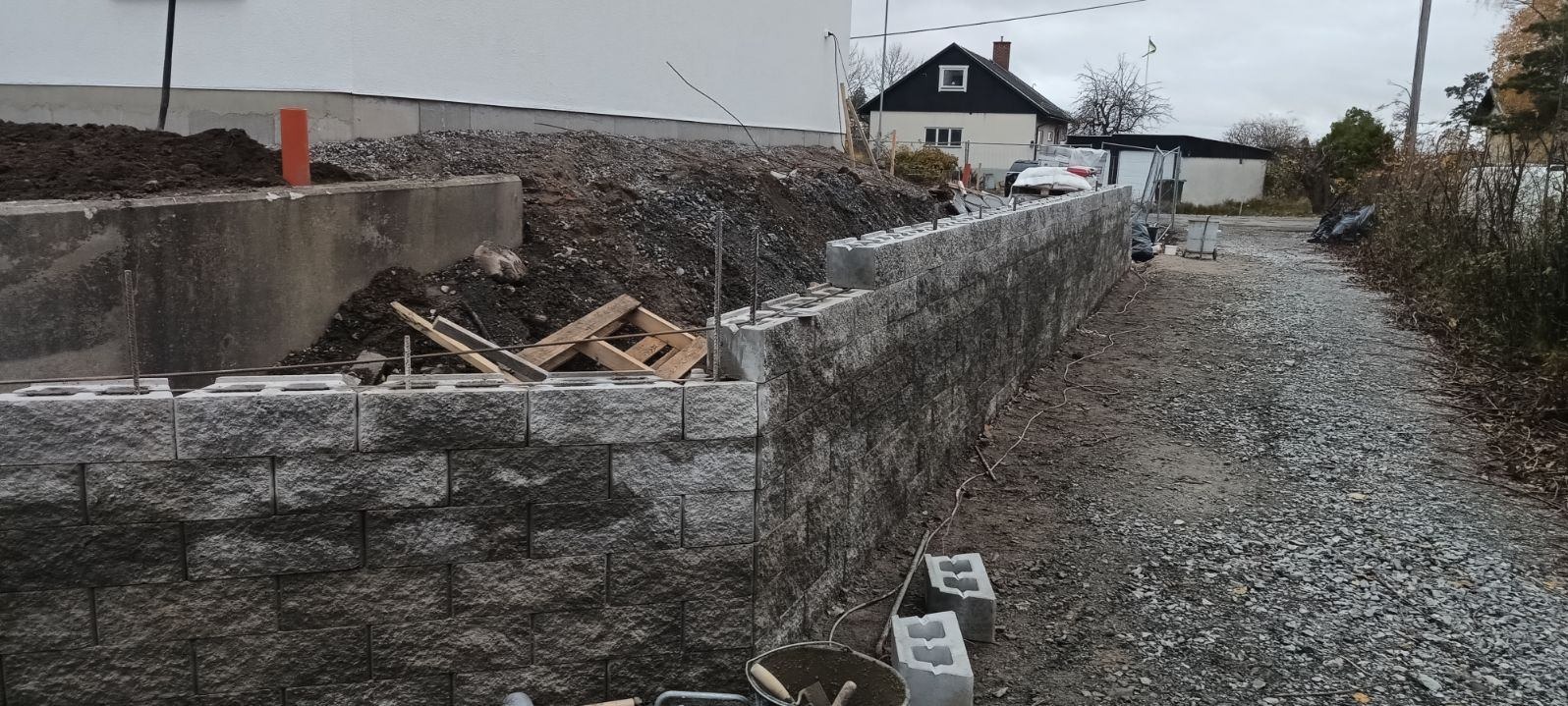 Wznoszenie muru oporowego z szarej kostki betonowej, widoczne zbrojenie, ziemia za murem i narzędzia murarskie w wiadrze u dołu kadru, w tle budynek mieszkalny.
