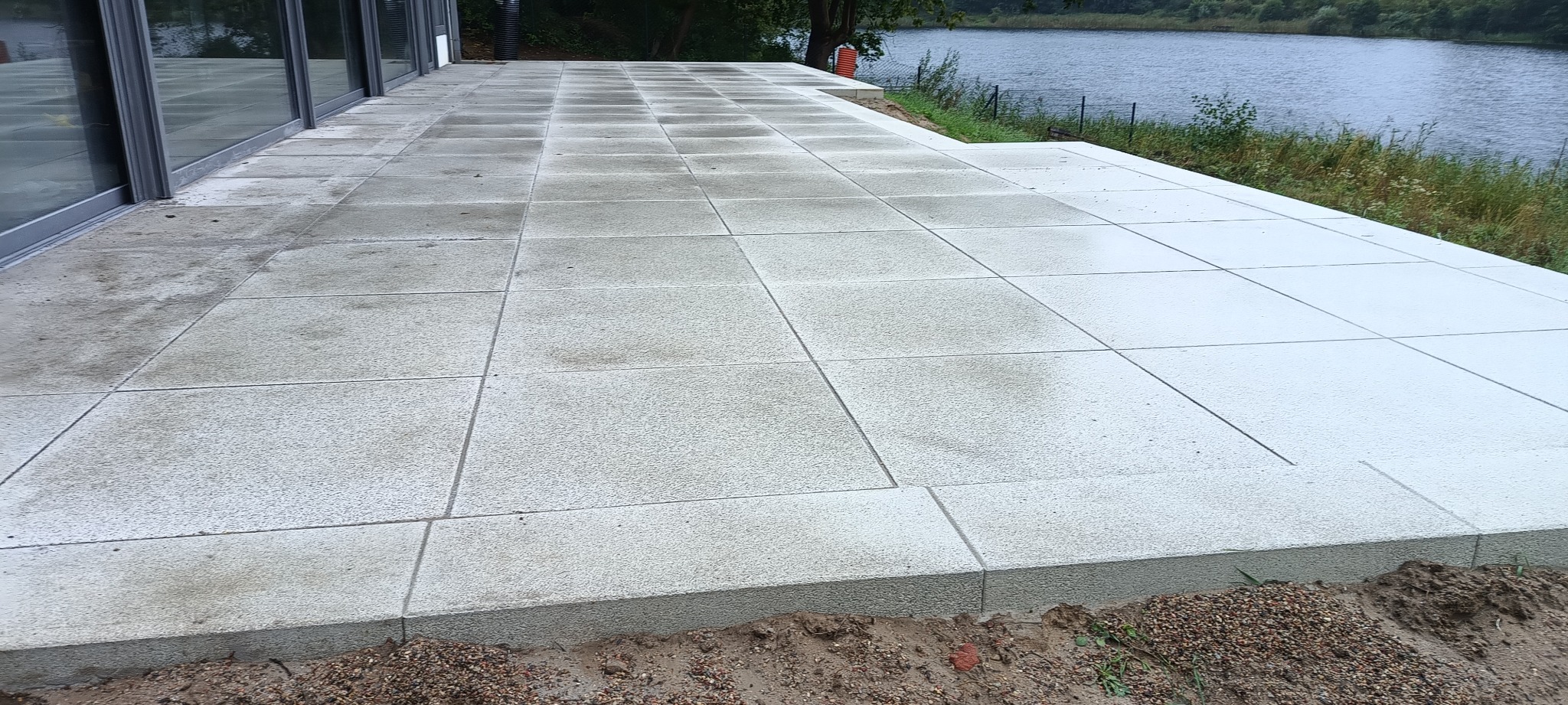 Nowoczesny taras z betonowych płyt w odcieniach szarości, z widokiem na jezioro w tle, świeżo obsypany ziemią.