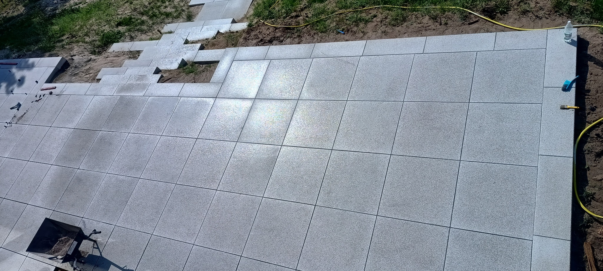 Szare płyty tarasowe ułożone w regularny wzór, częściowo na podłożu gruntowym, z widocznymi narzędziami i wózkiem wypełnionym piaskiem.