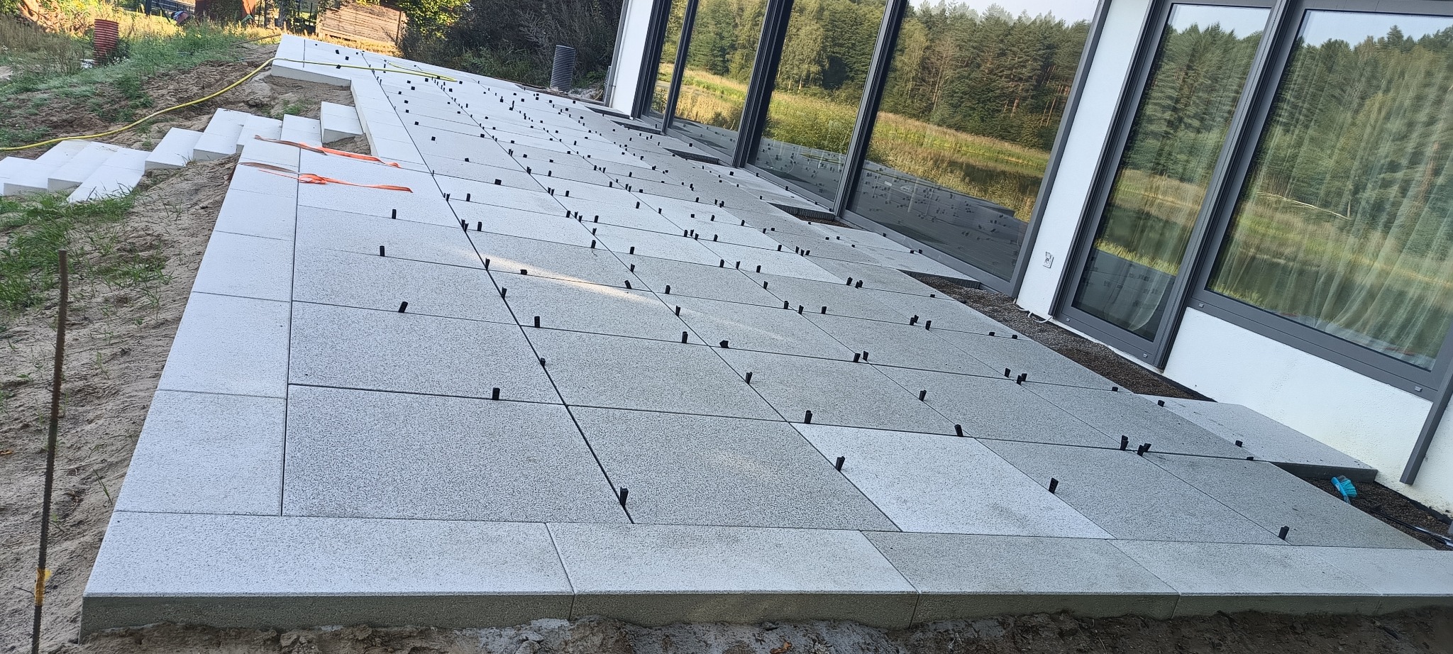 Montaż tarasu z płyt betonowych na zewnątrz budynku z dużymi oknami, widoczne czarne elementy poziomujące i schody z białych bloków styropianowych.