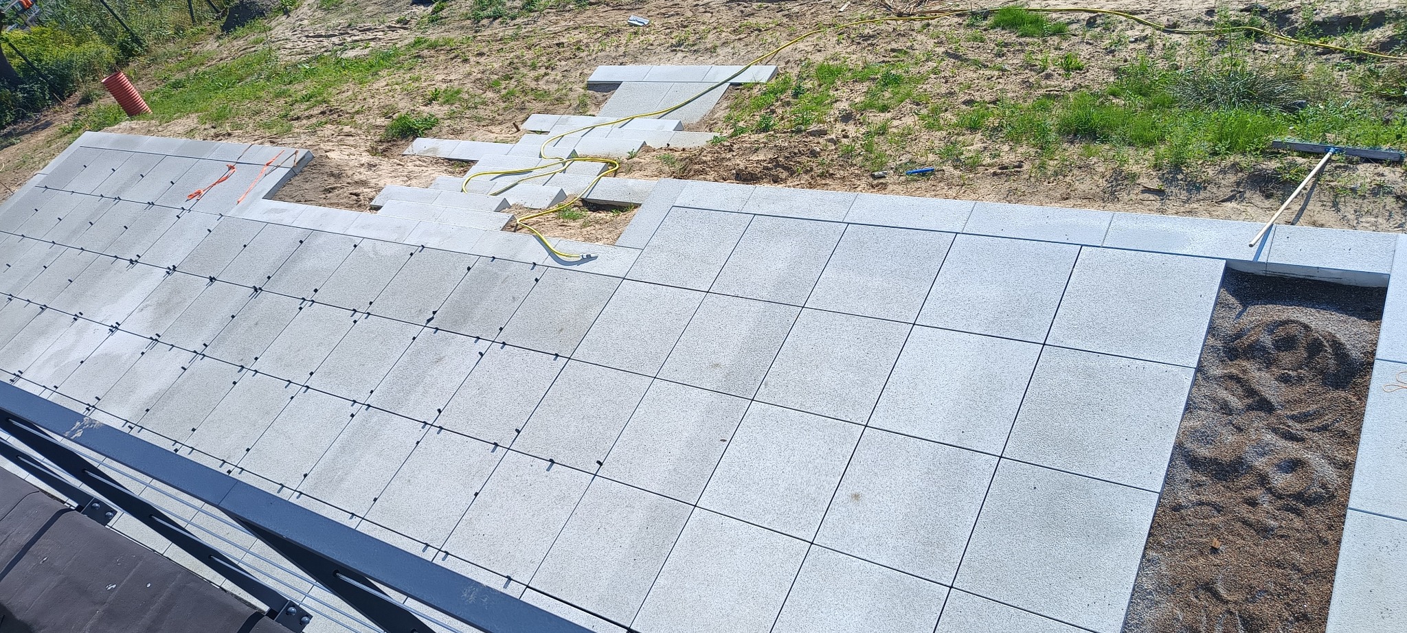 Układanie szarej kostki brukowej na piasku z użyciem sznurka i poziomicy, widoczne elementy mocujące między kostkami, żółty wąż ogrodowy i częściowo ułożone schody z kostki.