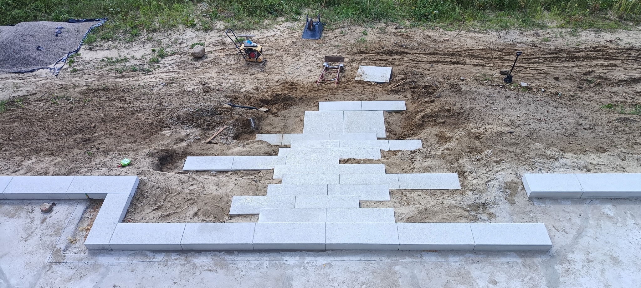 Wyrównany teren pod budowę tarasu z betonowych płyt, widoczne narzędzia i ubijarka w tle.