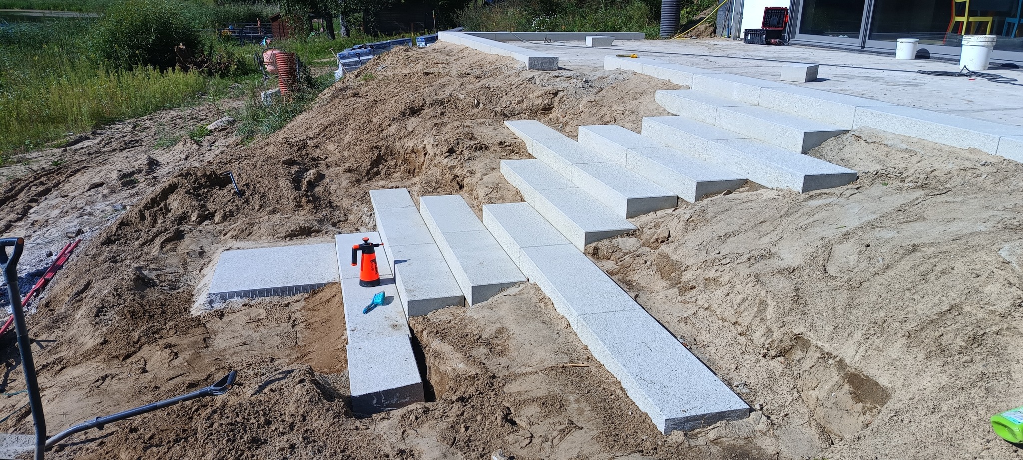 Budowa schodów zewnętrznych z betonowych elementów, widoczne narzędzia i piasek.