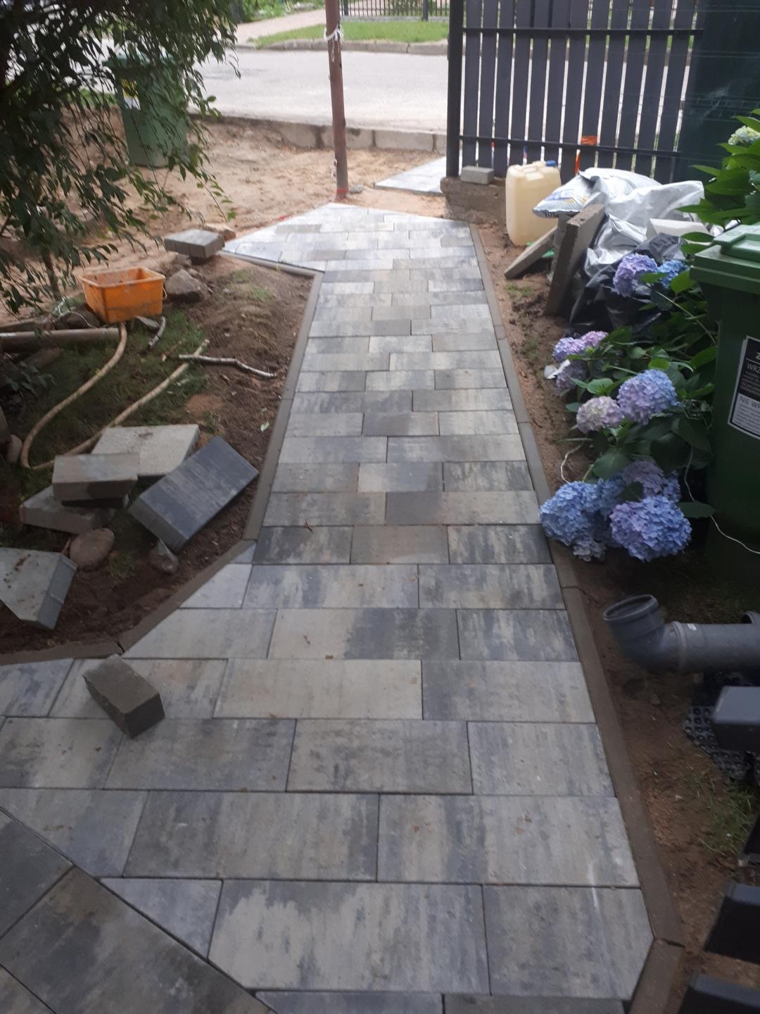 Ścieżka z szarej kostki brukowej w trakcie układania, widoczne niedokończone fragmenty i materiały budowlane oraz hortensje w tle.