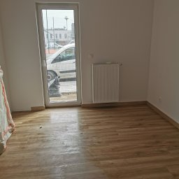 Farmont Gabriel Faryna - Pusty pokój po remoncie z panelami podłogowymi w odcieniach brązu, grzejnikiem na ścianie i widokiem na ulicę przez okno, z workiem gruzu za oknem.