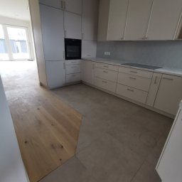 Farmont Gabriel Faryna - Jasna kuchnia z zabudową meblową, drewnianą podłogą przechodzącą w szarą płytkę, widoczny fragment salonu z oknami balkonowymi.