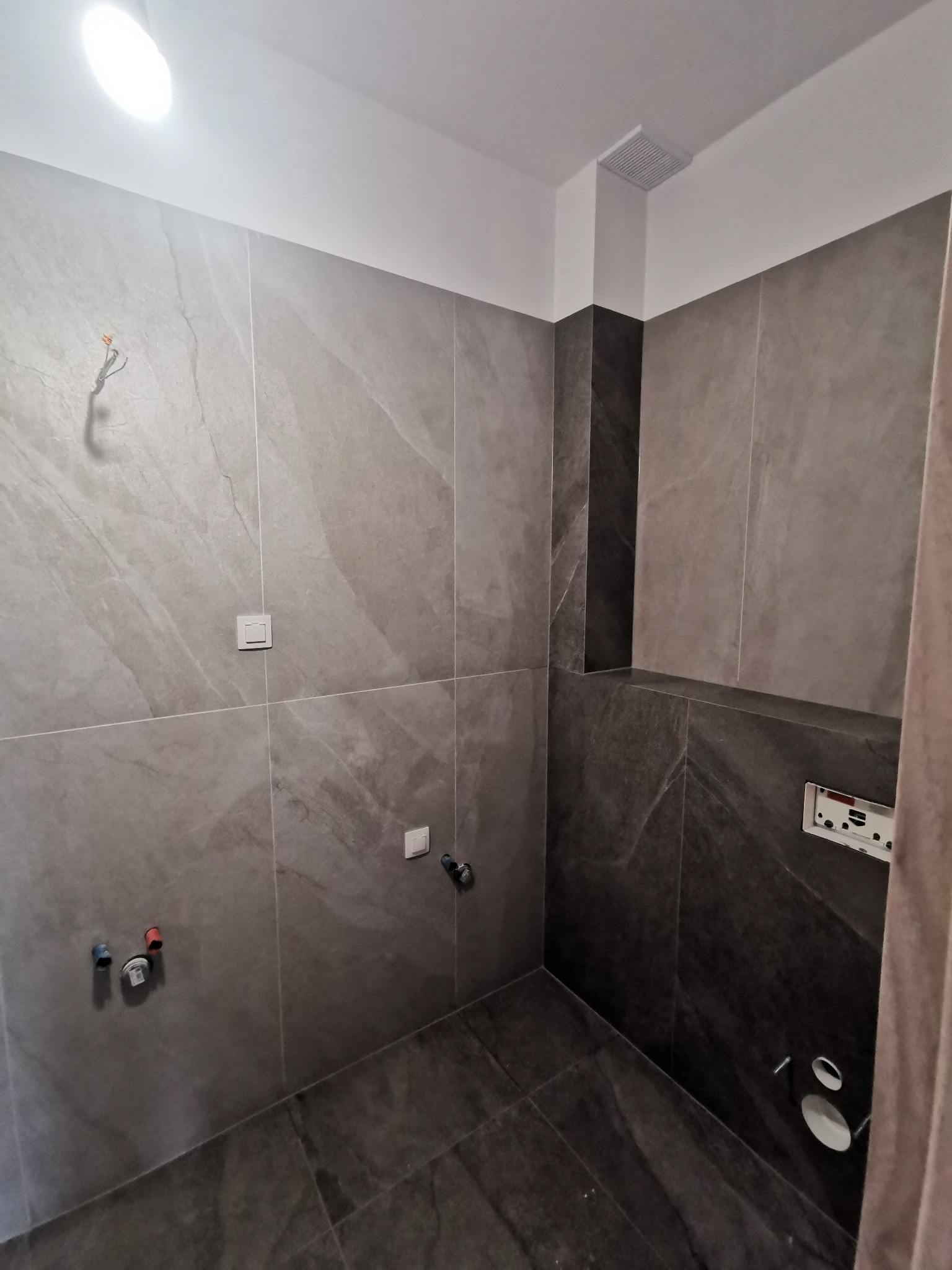 Wnętrze łazienki w trakcie wykańczania: szare płytki imitujące kamień na ścianach i podłodze, widoczne wystające kable elektryczne i rury, brak armatury, surowy stan.