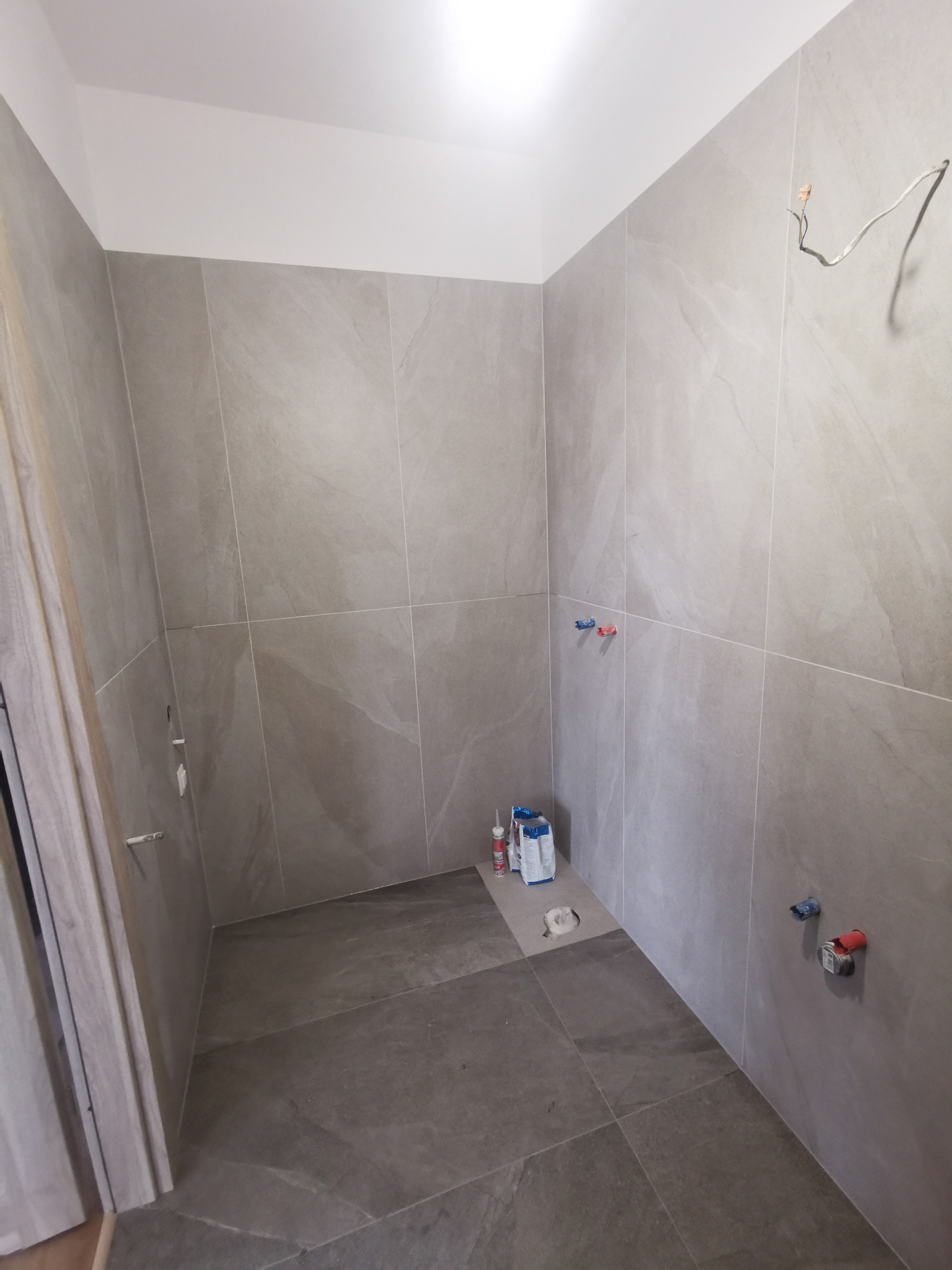 Łazienka w trakcie wykończenia: szare płytki podłogowe i ścienne, widoczne przyłącza hydrauliczne, puszka elektryczna z wystającym kablem oraz opakowania po materiałach budowlanych. Brak armatury...